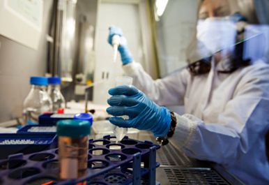 Picture5 | drmanishamehta.com Scientist in a lab coat wearing a mask and blue gloves uses a pipette over a rack of test tubes in a laboratory setup.