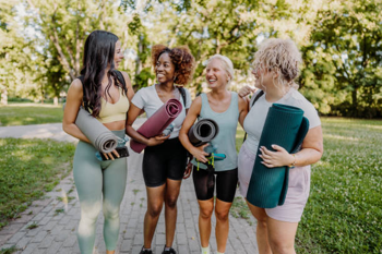 Four women in athletic wear stand on a park path holding rolled yoga mats, smiling and chatting.